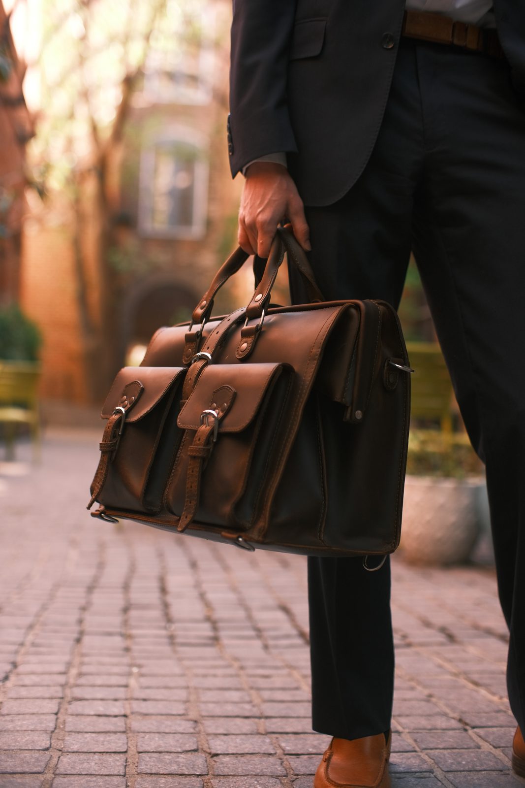 A brown leather briefcase with brass buckles and a shoulder strap rests on a wooden surface.