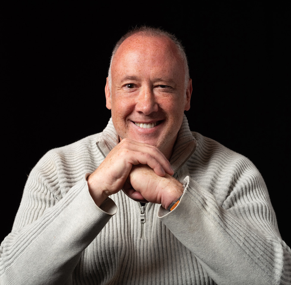Dan Jahn in a cream quarter-zip sweater, smiling with his hands folded under his chin against a black background